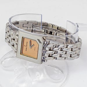 May include: A silver-toned Daniel Hechter watch with a square face and a linked bracelet. The watch face is peach-colored with black hands and the text "Daniel Hechter Paris". The watch is displayed on a clear plastic stand.