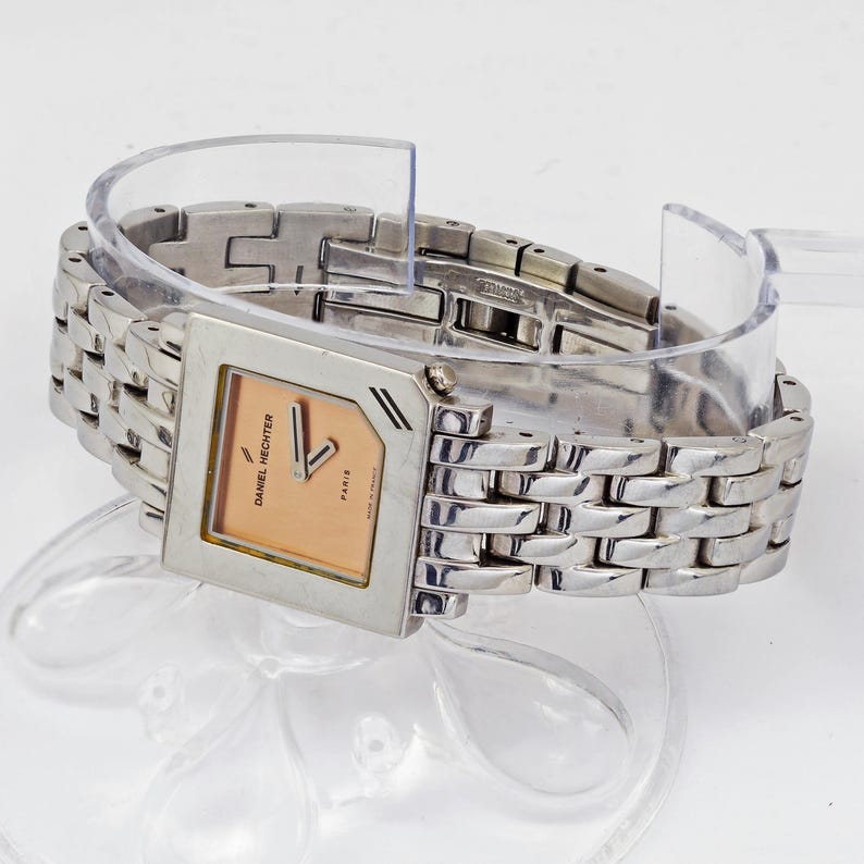 May include: A silver-toned Daniel Hechter watch with a square face and a linked bracelet. The watch face is peach-colored with black hands and the text "Daniel Hechter Paris". The watch is displayed on a clear plastic stand.