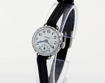 May include: A vintage wristwatch with a round silver-tone case featuring a textured bezel, a white dial with black Arabic numerals, blue hands, and a small sub-dial. The watch is attached to a thin black strap with visible stitching along the edges.