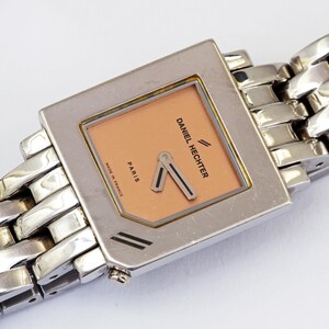 May include: A silver-toned Daniel Hechter wristwatch with a square face and a peach-colored dial. The watch features a silver link band and the words "Daniel Hechter" and "Paris" are visible on the face. The watch is marked "Made in France".