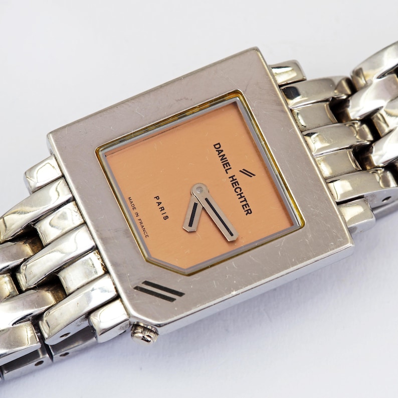 May include: A silver-toned Daniel Hechter wristwatch with a square face and a peach-colored dial. The watch features a silver link band and the words "Daniel Hechter" and "Paris" are visible on the face. The watch is marked "Made in France".