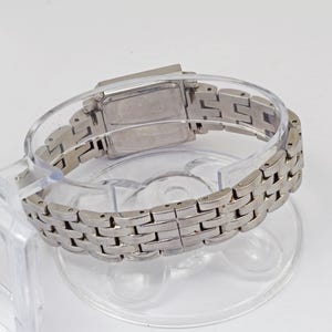May include: A silver-toned wristwatch with a rectangular face and a linked band. The watch is displayed on a clear plastic stand, showcasing its design and metallic finish. The band is made of small, rectangular links.