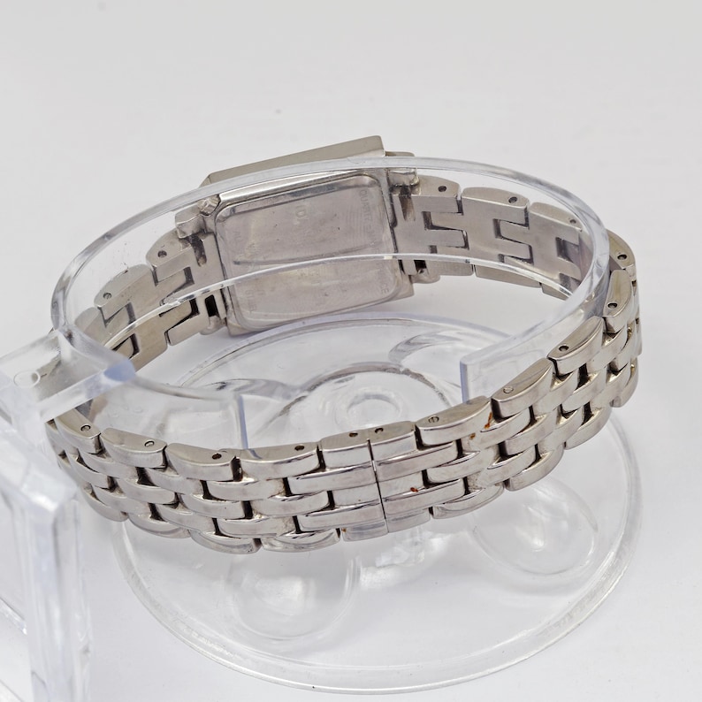 May include: A silver-toned wristwatch with a rectangular face and a linked band. The watch is displayed on a clear plastic stand, showcasing its design and metallic finish. The band is made of small, rectangular links.