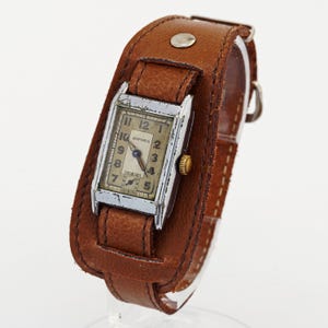 Könnte beinhalten: Eine rechteckige Bifora-Armbanduhr mit silberfarbenem Metallgehäuse und einem verwitterten cremefarbenen Zifferblatt mit schwarzen Ziffern. Die Uhr ist an einem breiten, braunen Lederarmband mit einem silbernen Metallniet und einer Dornschließe befestigt.