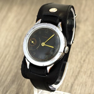 May include: Vintage wristwatch with a black face, silver-tone bezel, and yellow hands. The watch has a black leather cuff-style band. The watch face displays the numbers 1 through 12. The watch is a classic timepiece.