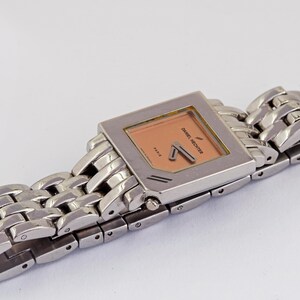 May include: A silver-toned Daniel Hechter watch with a rectangular peach-colored face and matching hands. The watch has a linked metal band. The watch face has the brand name and the word "Paris" printed on it.