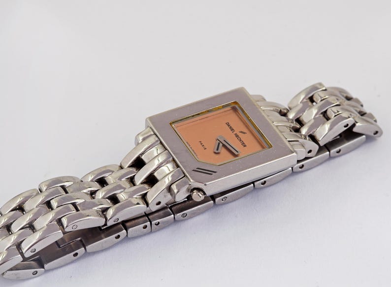 May include: A silver-toned Daniel Hechter watch with a rectangular peach-colored face and matching hands. The watch has a linked metal band. The watch face has the brand name and the word "Paris" printed on it.