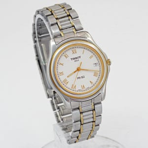 May include: A silver and gold Tissot PR 50 wristwatch with a white face and gold hands and accents. The watch features Roman numeral hour markers and a date display at the 3 o'clock position. The watch band is silver and gold.