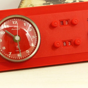 Op de afbeelding: Een vintage rode wekker met een ronde wijzerplaat en een rechthoekige basis. De wijzerplaat heeft witte cijfers en wijzers, en de basis heeft draaiknoppen en een schakelaar. De klok heeft een retro-ontwerp.