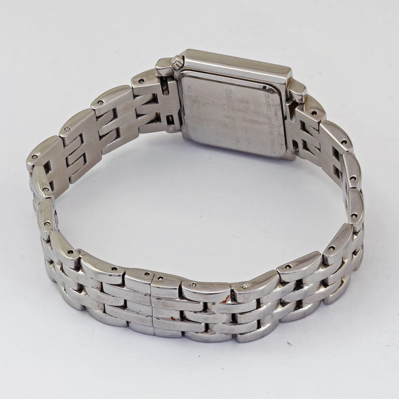 May include: A silver-toned wristwatch with a rectangular face and a linked band. The watch has a metallic sheen and a classic design. The band is composed of multiple small, rectangular links. The watch is shown against a white background.