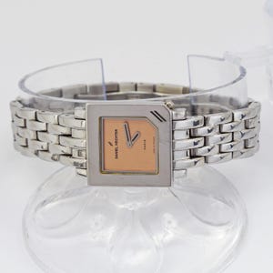 May include: A Daniel Hechter Paris wristwatch with a silver-toned rectangular case and band. The watch face is peach-colored with silver hands and markers. The band is made of interlocking links.