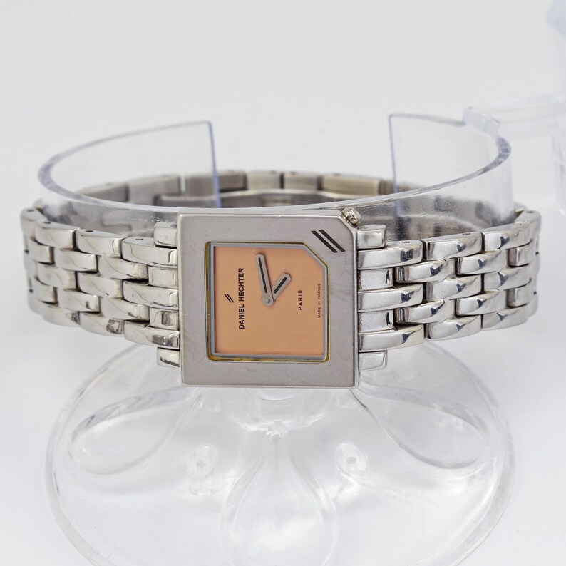 May include: A Daniel Hechter Paris wristwatch with a silver-toned rectangular case and band. The watch face is peach-colored with silver hands and markers. The band is made of interlocking links.
