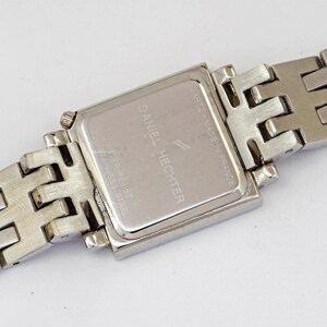 May include: Close-up of a silver Daniel Hechter quartz watch. The watch has a rectangular face and a stainless steel band. The back of the watch is visible, with the brand name and other details printed on it. The watch is water resistant.