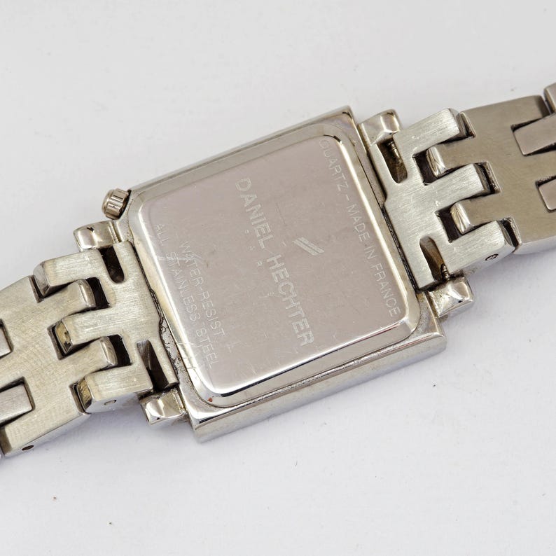 May include: Close-up of a silver Daniel Hechter quartz watch. The watch has a rectangular face and a stainless steel band. The back of the watch is visible, with the brand name and other details printed on it. The watch is water resistant.
