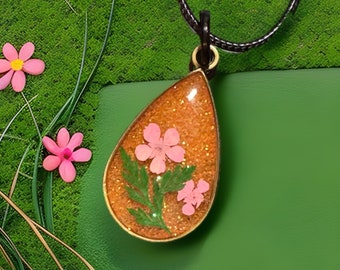May include: A teardrop-shaped pendant necklace with a gold-colored frame. The pendant contains dried pink flowers and green leaves set in a sparkly, amber-colored resin. The necklace hangs on a black cord.