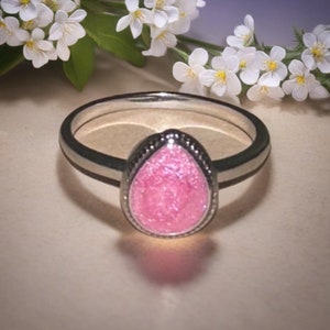 May include: A silver ring with a pear-shaped, pink, glittery stone. The ring has a dark band with a silver stripe on each side. The stone is framed by a beaded silver border. White flowers are in the background.