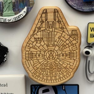 May include: A wooden magnet shaped like the Millennium Falcon spaceship from Star Wars. The magnet is light brown with detailed engraved lines. Other magnets include the Statue of Liberty and a paperclip with googly eyes.