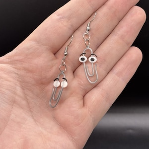 Puede incluir: Un par de pendientes originales con dise&ntilde;os de clips plateados con ojos saltones. Los pendientes tienen un cierre de gancho sencillo y se muestran sobre un fondo neutro.