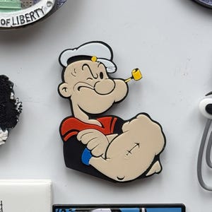 Puede incluir: Una imagen de dibujos animados de Popeye el Marino con su característico traje de marinero rojo y azul, un gorro de marinero blanco y una pipa. Está flexionando el bíceps y tiene una expresión decidida en su rostro.