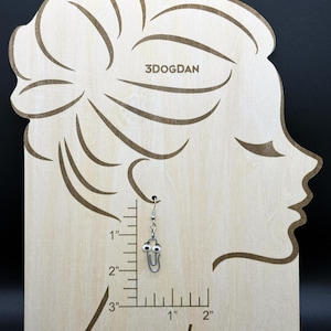 Puede incluir: Un pendiente de clip plateado con ojos saltones cuelga de una pantalla de madera. La pantalla tiene la forma de un perfil de mujer y tiene el texto "3DOGDAN" grabado. El pendiente mide aproximadamente 2,5 cm de largo.