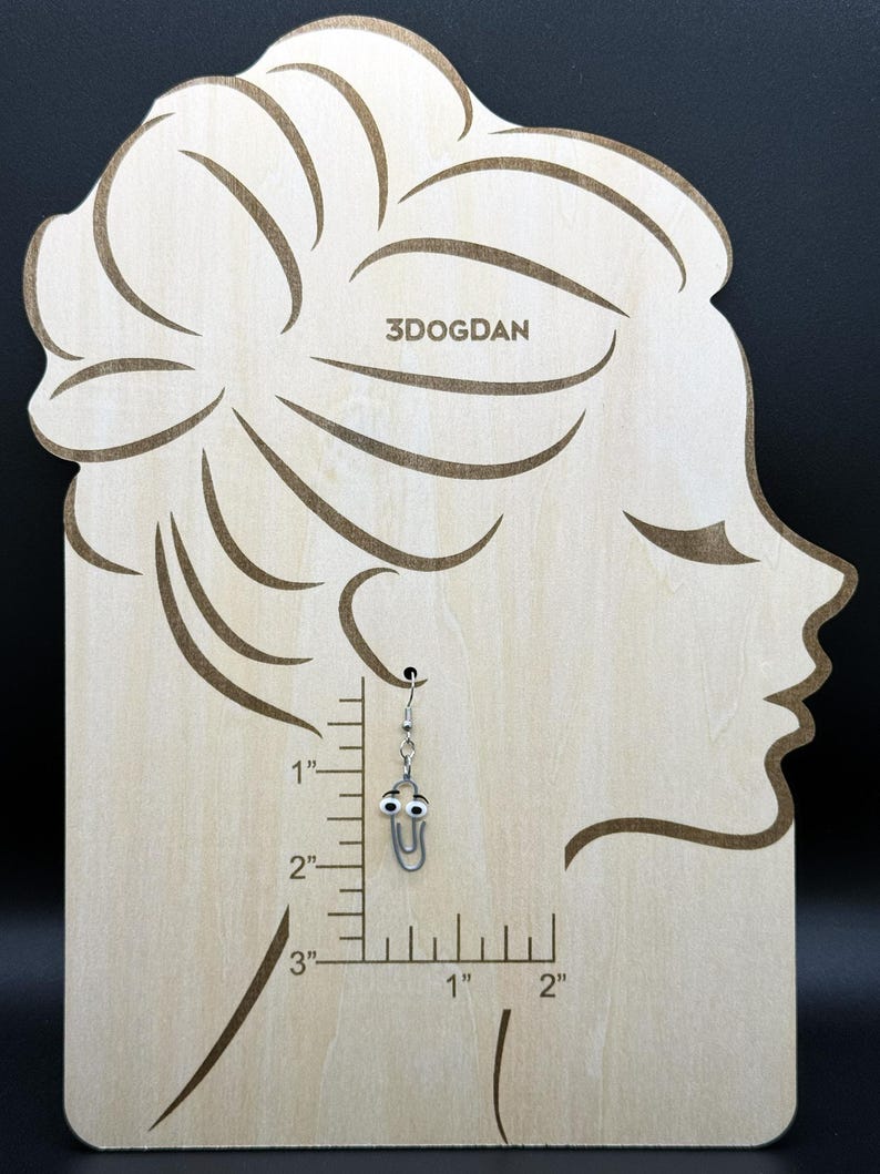 Puede incluir: Un pendiente de clip plateado con ojos saltones cuelga de una pantalla de madera. La pantalla tiene la forma de un perfil de mujer y tiene el texto "3DOGDAN" grabado. El pendiente mide aproximadamente 2,5 cm de largo.