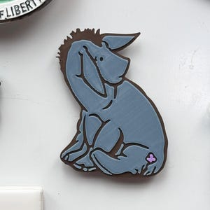 May include: A blue 3D printed figurine of Eeyore the donkey from Winnie the Pooh, sitting with his head in his hand. He has a small purple flower on his back.