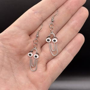 Puede incluir: Un par de pendientes originales con un dise&ntilde;o de clip plateado con ojos saltones. Los pendientes tienen un cierre de gancho sencillo y se sostienen en una mano sobre un fondo oscuro. Los ojos son blancos con pupilas negras y cejas negras.