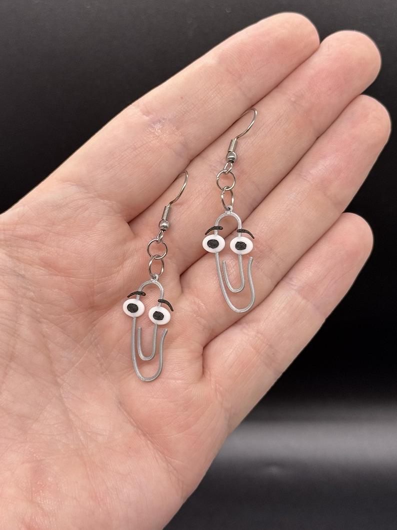 Puede incluir: Un par de pendientes originales con un dise&ntilde;o de clip plateado con ojos saltones. Los pendientes tienen un cierre de gancho sencillo y se sostienen en una mano sobre un fondo oscuro. Los ojos son blancos con pupilas negras y cejas negras.