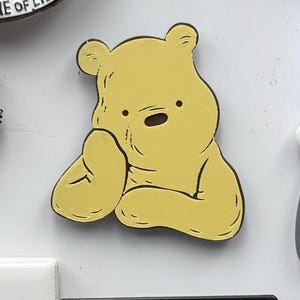 May include: A yellow cartoon bear-shaped magnet with a black outline. The bear is in a thinking pose, with its hand on its cheek. The magnet is on a white surface, with other magnets and a paperclip visible.