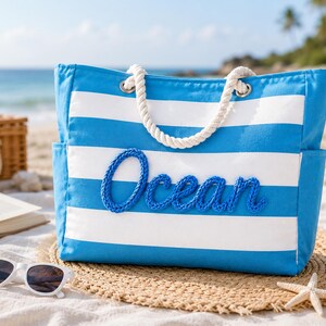 May include: A blue and white striped canvas tote bag featuring the word Ocean in raised blue rope lettering on the front. The bag has thick white twisted rope handles attached with silver metal grommets and side pockets on both ends.