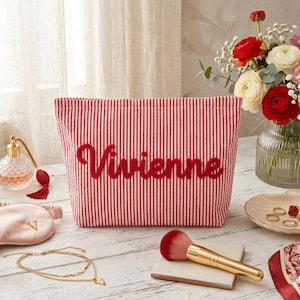 Può includere: Un beauty case a righe rosse e bianche con il nome "Vivienne" ricamato in rosso. L'immagine include anche un flacone di profumo, gioielli, un pennello per il trucco e un bouquet di fiori.