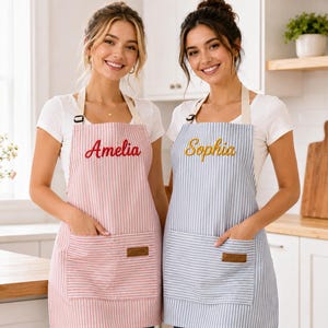 Op de afbeelding: Twee gestreepte schorten met verstelbare nekbanden en voorzakken. Het ene schort is roze met witte strepen en de naam Amelia in het rood geborduurd. Het andere is blauw met witte strepen en de naam Sophia in het goudgeel geborduurd.
