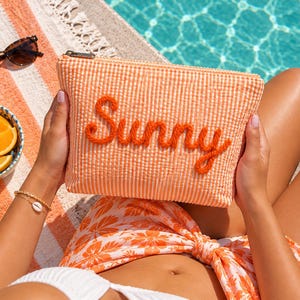 Könnte beinhalten: Eine rechteckige Reißverschlusstasche aus orange-weißem Vichy-Stoff. Das Wort Sunny ist auf der Vorderseite mit dickem, strukturiertem orangefarbenem Garn aufgestickt. Die Tasche hat einen silberfarbenen Metallreißverschluss oben rechts.