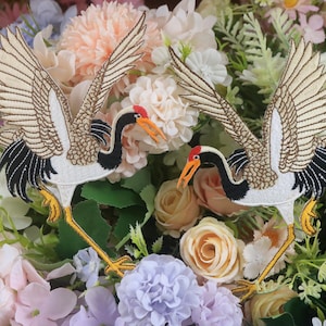 May include: Two embroidered crane patches with outstretched wings, featuring beige, black, white, red, and yellow details. The patches are set against a backdrop of colorful flowers, showcasing a nature-inspired theme.