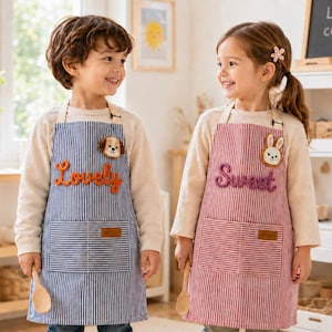 May include: Two children's aprons, one blue and white striped with a dog patch and the word Lovely, the other red and white striped with a rabbit patch and the word Sweet. Both feature a front pocket with a small rectangular label and neck straps.