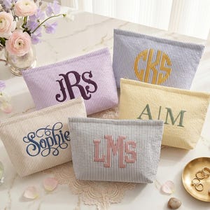May include: Five rectangular zippered pouches made of seersucker fabric with vertical stripes in various pastel colors. Each pouch features custom embroidery, including monograms like JRS, CKS, LMS, AM, and the name Sophie in dark thread.