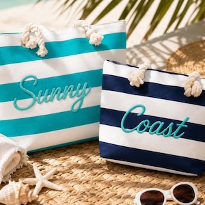 May include: Two tote bags with rope handles. The larger bag features horizontal teal and white stripes with the word Sunny embroidered in teal. The smaller bag has horizontal navy blue and white stripes with the word Coast embroidered in teal.