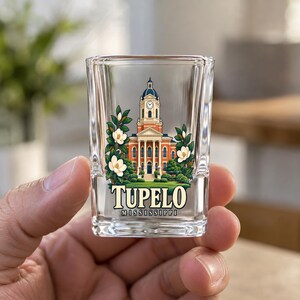 May include: A clear glass shot glass featuring a colorful illustration of a brick courthouse with a clock tower, surrounded by white magnolia flowers and green leaves. The text TUPELO MISSISSIPPI is printed in bold lettering below the building.