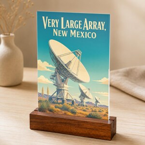 May include: A rectangular print featuring a graphic illustration of the Very Large Array in New Mexico, with large satellite dishes in a desert landscape under a blue sky, held upright by a dark brown rectangular wooden block stand.
