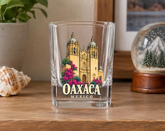 May include: A clear square shot glass featuring a colorful illustration of a cathedral with two towers and pink bougainvillea flowers. The text OAXACA MEXICO is printed in black lettering below the building on the front of the glass.