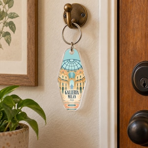 May include: A motel-style keychain featuring a light blue and tan illustration of the Galleria Vittorio Emanuele II in Milan, Italy. The keychain has a rounded, elongated shape with a metal ring attached to a brass-colored wall hook.