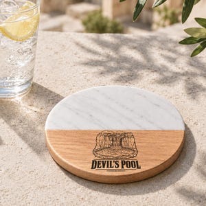 May include: A circular coaster featuring a split design with white marble on the top half and light brown wood on the bottom half. The wooden section displays a black engraved illustration of a waterfall and pool with the text DEVIL'S POOL below.
