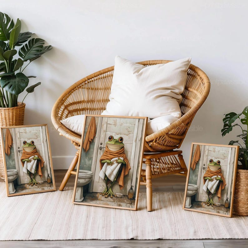 May include: Three framed art prints featuring a whimsical frog character in a bathroom setting. The frog is depicted wearing a towel and robe, standing in front of a door. The prints are displayed near a wicker chair and potted plants.