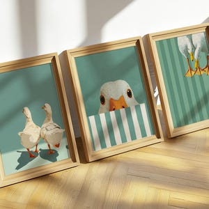 Op de afbeelding: Drie ingelijste kunstprints met grillige ganzenillustraties. De prints hebben een turquoise achtergrond met de ganzen in verschillende poses. De frames zijn gemaakt van lichtgekleurd hout, wat een natuurlijke touch aan het kunstwerk toevoegt.