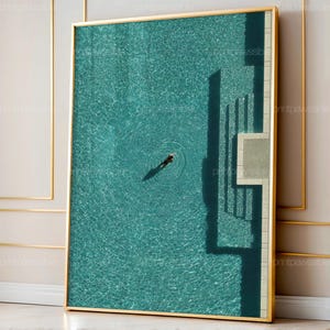 Puede incluir: Una impresión artística enmarcada que presenta una vista aérea de una piscina turquesa con un nadador. La superficie de la piscina tiene ondulaciones y el marco es dorado. La obra captura el juego de luces y sombras.