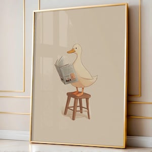 May include: A framed art print features a cartoon duck reading a newspaper while perched on a small wooden stool. The duck is white with an orange beak and feet. The background is a neutral beige color, and the frame is gold.