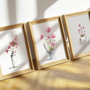 May include: Three framed watercolor art prints featuring pink flowers. One print shows flowers in a spray paint can, another in a toilet, and the third in a roll of toilet paper. The frames are made of light-colored wood.