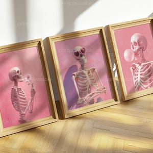 May include: Three framed art prints featuring skeleton illustrations. One skeleton holds a flower, another wears heart-shaped sunglasses, and the third blows a bubble. Each print has a pink background and a wooden frame.