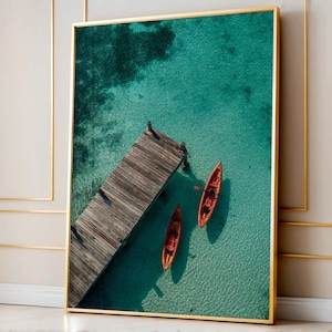 Puede incluir: Impresión enmarcada que muestra una vista aérea de un muelle de madera que se extiende hacia aguas turquesas claras. Dos kayaks naranjas están cerca del muelle. La impresión está en un marco dorado, creando una escena costera serena.