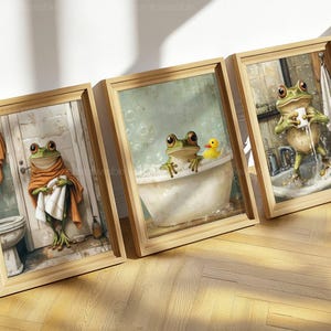 May include: Three framed art prints featuring whimsical frog illustrations in a bathroom setting. One frog is wrapped in a towel, another in a bathtub with a rubber duck, and the third washing its face at a sink. The frames are light brown.